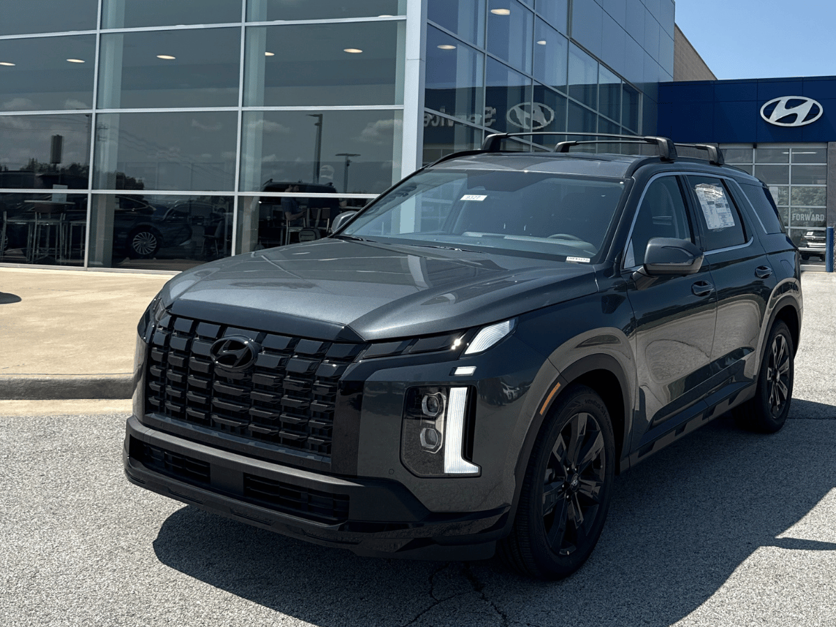 Front angle of a 2025 Hyundai Palisade SUV in dark gray parked outside Crain Hyundai of Fayetteville’s showroom.